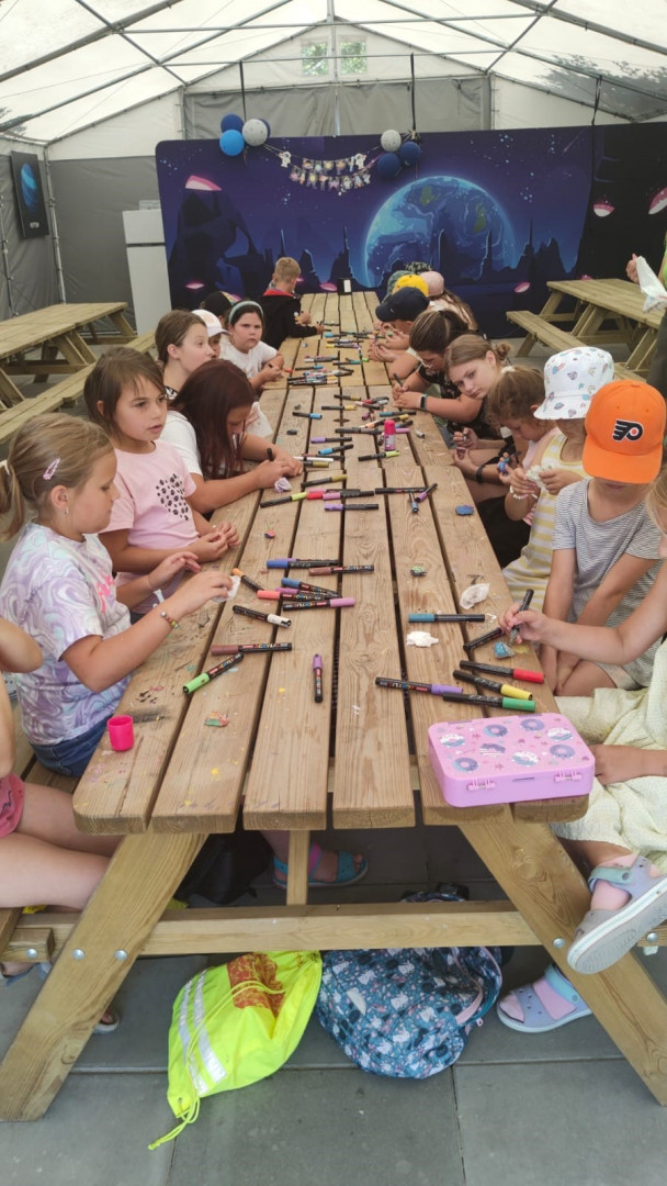 Wokół długiego stołu siedzi grupa dzieci w wieku szkolnym. Na stole leżą kredki i kartki do rysowani